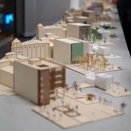 A series of architectural models on a table in the urban laboratory at RheinMain University of Applied Sciences and Arts. These are the thesis projects of bachelor's and master's students. The models are mainly made of wood and show different shapes, sizes, and some trees. 