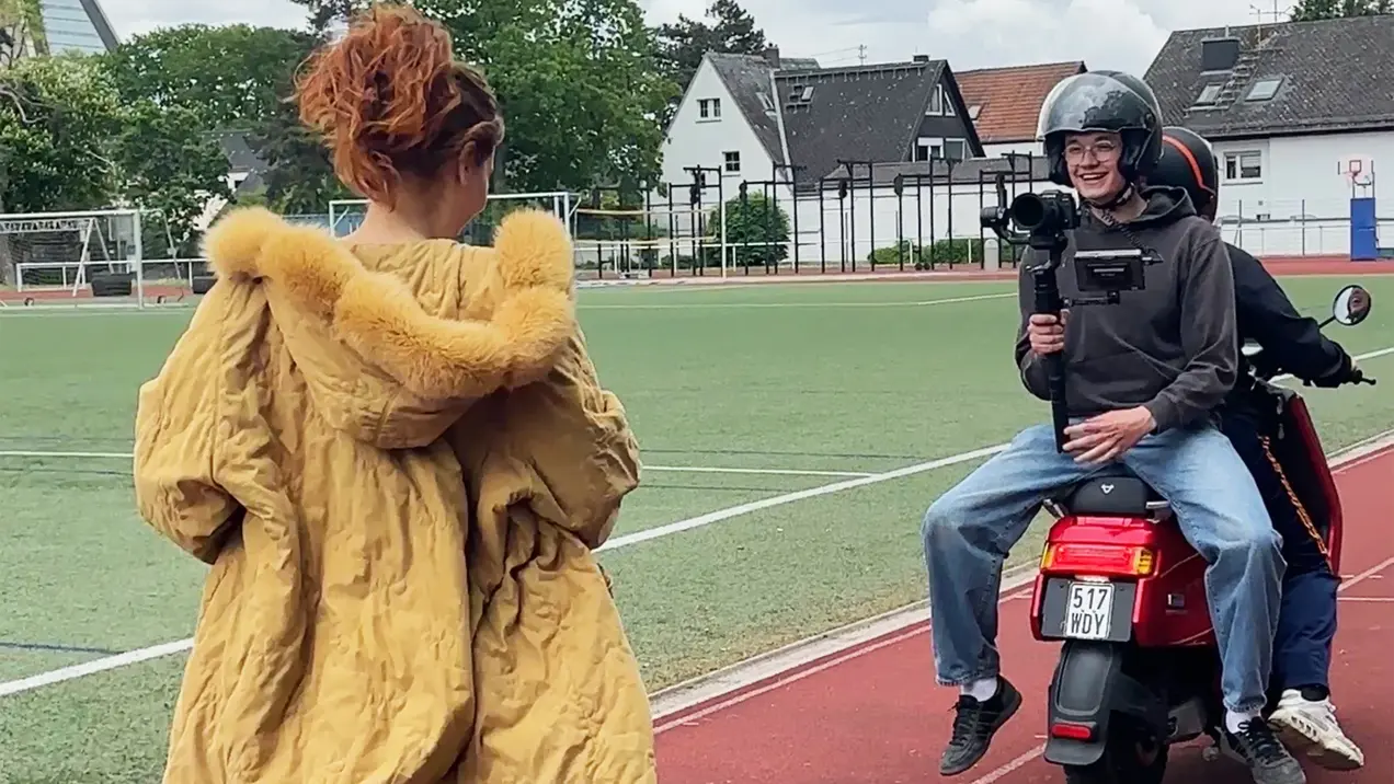 Eine Person in einem auffälligen gelben Mantel steht auf einem Sportplatz und schaut in die Richtung eines jungen Mannes, der auf einem motorisierten Roller sitzt. Er hält eine Kamera und lächelt, während er sie filmt. Der Hintergrund zeigt Fußballfelder und Gebäude.