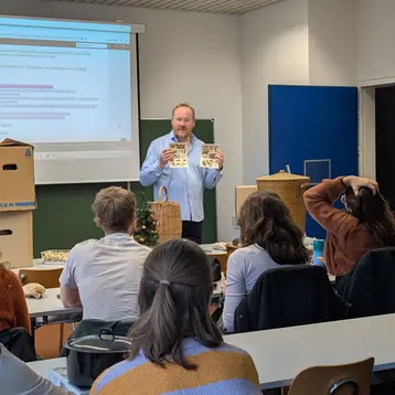 Ein Referent steht in einem Seminarraum und hält zwei Plakate in den Händen. Im Hintergrund ist eine Präsentation zu sehen, im Vordergrund Studierende, die aufmerksam zuhören. Es handelt sich um ein Seminar zum Thema ressourcenschonendes Bauen und ressourcenschonendes Weihnachten.