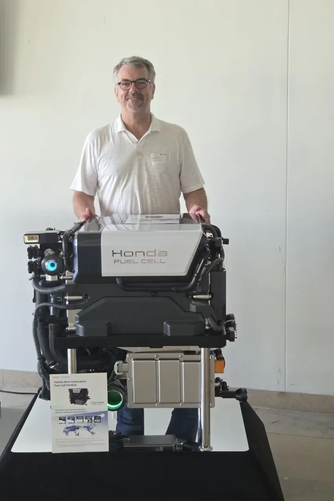 Ein Mann steht neben einem Honda Brennstoffzellenmotor, der auf einem Tisch präsentiert wird. Der Motor ist sichtbar, mit klaren Kennzeichnungen. Im Vordergrund liegt eine Informationsbroschüre mit technischen Details. Der Hintergrund ist hell und neutral.