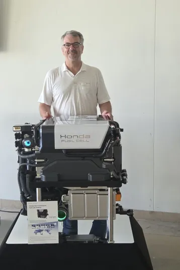 Ein Mann steht neben einem Honda Brennstoffzellenmotor, der auf einem Tisch präsentiert wird. Der Motor ist sichtbar, mit klaren Kennzeichnungen. Im Vordergrund liegt eine Informationsbroschüre mit technischen Details. Der Hintergrund ist hell und neutral.