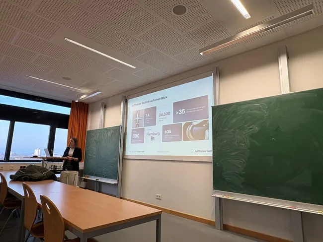 Eine Person steht vor einer Präsentation in einem Hörsaal. Die Folie zeigt verschiedene Zahlen und Statistiken in Bezug auf Lufthansa Technik, darunter Daten zu Standorten und Mitarbeiterzahlen. Zwei grüne Tafeln sind im Hintergrund sichtbar.