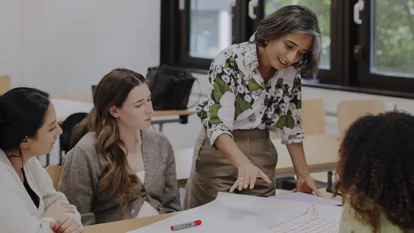 Man sieht drei Studierende im Vordergrund, die gemeinsam an einem Projekt arbeiten und eine Professorin, die gerade etwas erklärt. Vor Ihnen auf dem Tisch liegen große Blätter mit aufgeschriebenen Ideen. Im Hintergrund sieht man große Fenster.