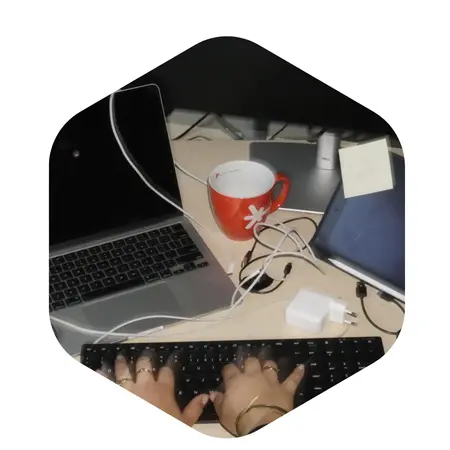 Eine Person tippt auf einer Tastatur vor einem Laptop. Neben dem Laptop steht eine rote Tasse und ein Tablet. Kabel sind auf dem Schreibtisch sichtbar, und ein Zettel mit einer Notiz liegt ebenfalls daneben.