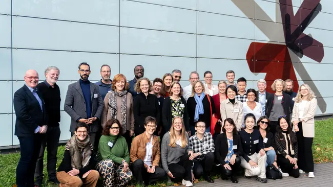 Eine Gruppe von etwa 40 Personen steht im Freien vor einer modernen Wand und lächelt in die Kamera. Die Teilnehmer sind divers gekleidet, und im Hintergrund ist ein großes, künstlerisches Wandbild zu sehen. Die Szene vermittelt ein Gefühl von Gemeinschaft und Zusammenarbeit.