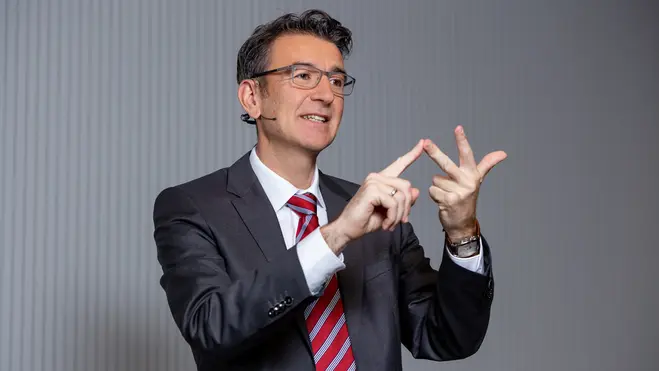 Ein Mann in einem Anzug mit Krawatte steht im Vordergrund und gestikuliert mit beiden Händen. Er zeigt mit seinen Fingern ein Konzept oder eine Idee. Der Hintergrund ist hell und schlicht. Der Mann wirkt engagiert und spricht möglicherweise zu einem Publikum.