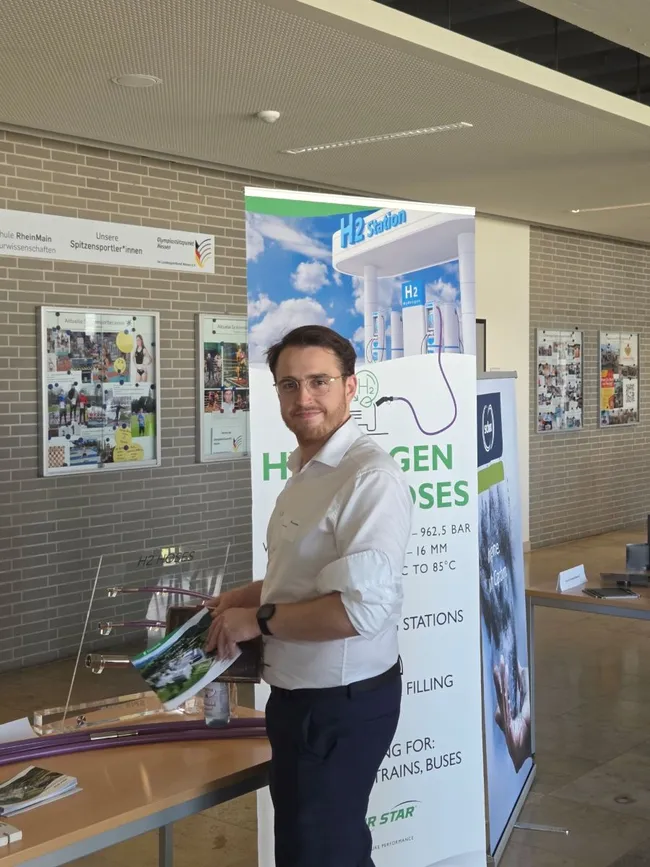 Ein Mann steht an einem Informationsstand zur Wasserstofftechnologie. Er hält Broschüren in der Hand und lächelt. Im Hintergrund ist ein Banner mit dem Schriftzug „Wasserstoff tanken“ zu sehen, das Informationen zu Wasserstofftankstellen bietet.