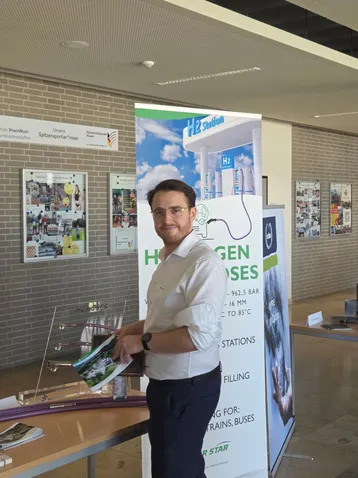 Ein Mann steht an einem Informationsstand zur Wasserstofftechnologie. Er hält Broschüren in der Hand und lächelt. Im Hintergrund ist ein Banner mit dem Schriftzug „Wasserstoff tanken“ zu sehen, das Informationen zu Wasserstofftankstellen bietet.