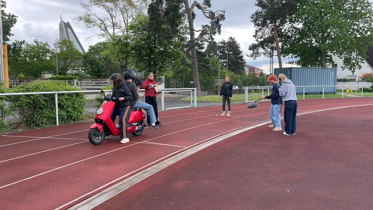 Fünf Personen stehen auf einer Laufbahn. Eine Person fährt auf einem roten Roller, während andere zuschauen und miteinander sprechen. Im Hintergrund sind Bäume und ein Gebäude sichtbar. Der Himmel ist bewölkt.