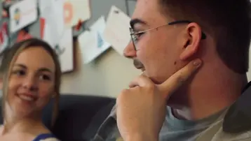 Zwei Personen sitzen auf einem Sofa und unterhalten sich. Eine Frau lächelt, während ein Mann nachdenklich mit dem Finger an seinem Kinn sitzt. Die Atmosphäre ist freundlich und entspannt.