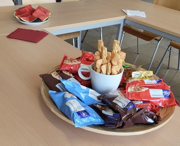 Eine große Holzschüssel steht auf einem Tisch, gefüllt mit verschiedenen Snacks: Keksen und kleinen Gebäcken. Ein paar Stücke Wassermelone liegen auf einem anderen Teller im Hintergrund. Die Atmosphäre wirkt einladend und gemütlich.