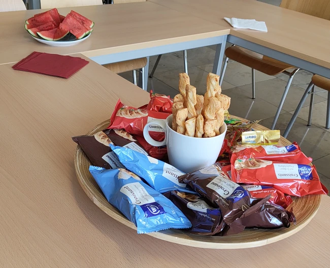 Eine große Holzschüssel steht auf einem Tisch, gefüllt mit verschiedenen Snacks: Keksen und kleinen Gebäcken. Ein paar Stücke Wassermelone liegen auf einem anderen Teller im Hintergrund. Die Atmosphäre wirkt einladend und gemütlich.
