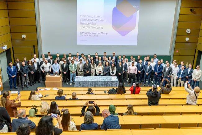 Gruppenfoto einer großen Gruppe von Menschen, die sich in einem Auditorium versammelt haben. Im Hintergrund befindet sich eine Präsentation mit der Aufschrift „Einladung zum gemeinsamen Gremiumfoto und Sektempfang“. Die Anwesenden lächeln und posieren, während einige Fotos machen.