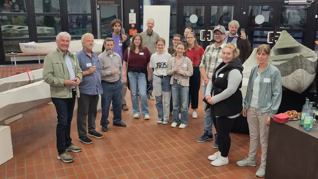 Eine Gruppe von etwa 20 Personen versammelt sich in einem Raum. Sie lächeln und halten Gläser in der Hand. Im Hintergrund sind einige Tisch- und Stuhlkonstruktionen sowie Dekorationsobjekte zu sehen. Die Atmosphäre wirkt freundlich und einladend.