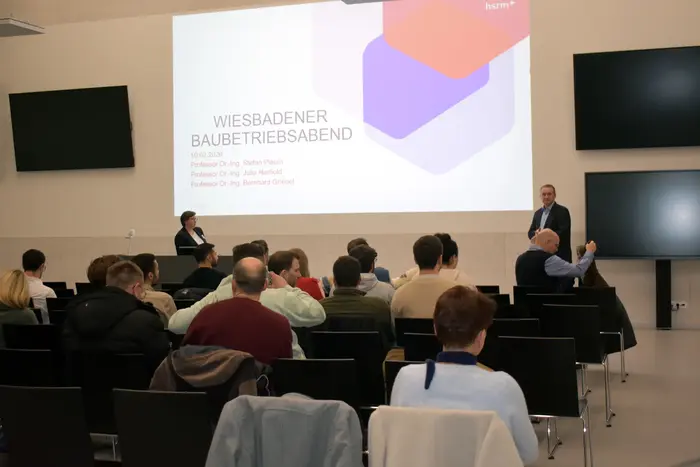 Prof. Julia Herhold und Prof. Bernhard Griebel begrüßen beim "Wiesbadener Baubetriebsabend" an der Hochschule RheinMain in einem Raum des neuen L-Gebäudes statt. Das Publikum sitzt in Reihen. Auf der Leinwand sind Informationen über das Event und die Referenten zu sehen.