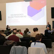 Prof. Julia Herhold und Prof. Bernhard Griebel begrüßen beim "Wiesbadener Baubetriebsabend" an der Hochschule RheinMain in einem Raum des neuen L-Gebäudes statt. Das Publikum sitzt in Reihen. Auf der Leinwand sind Informationen über das Event und die Referenten zu sehen.