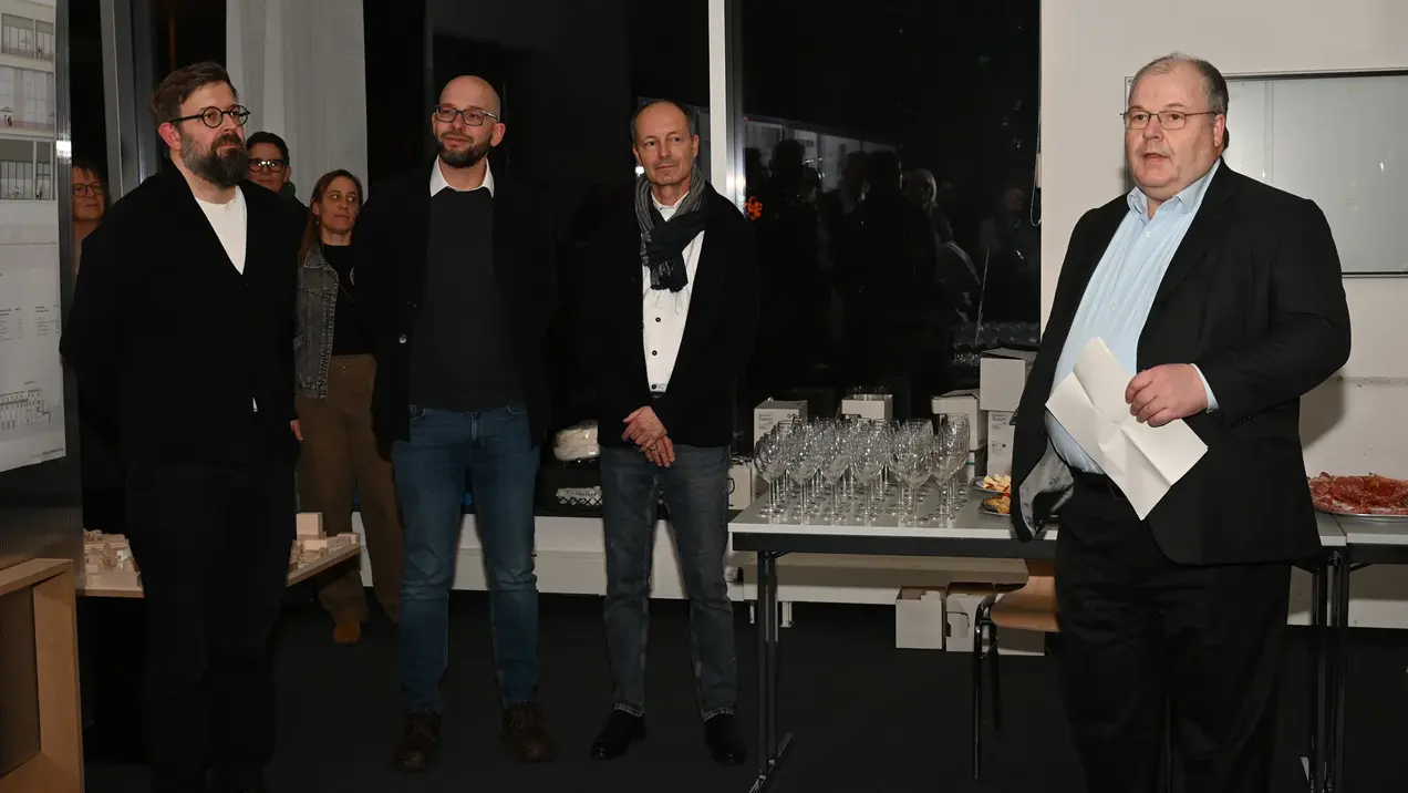 Eine Gruppe von vier Männern steht in einem Raum. Es handelt sich um Prof. Daniel Seiberts, Prof. Dr. Matthias Kowald, Prof. Dieter Müller und Prof. Dr. Andreas Brensing von der Hochschule RheinMain. Vizepräsident Prof. Dr. Brensing hält ein Papier in der Hand und spricht. Im Hintergrund sind Gläser und Snacks auf einem Tisch sichtbar. Die Atmosphäre scheint gesellig und formell zu sein.