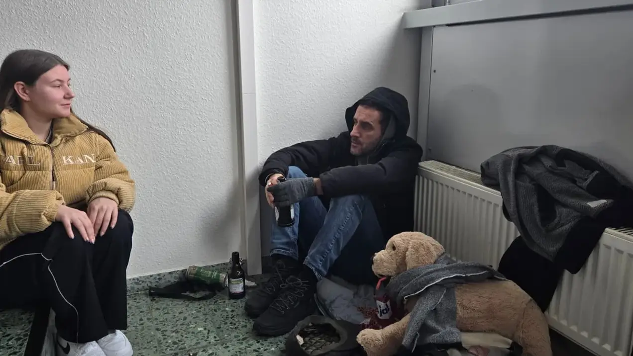Zwei Personen sitzen in einer Ecke eines Raumes. Eine Frau in einer gelben Jacke lächelt, während ein Mann in dunkler Kleidung ihr gegenüber sitzt. Neben ihnen liegt ein Plüschtier in Form eines Hundes und eine Flasche ist sichtbar.