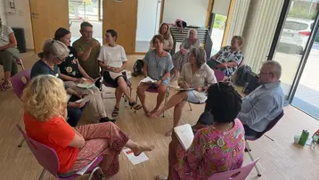 Eine Gruppe von etwa 15 Personen sitzt in einem hellen Raum auf Stühlen. Einige Menschen sprechen miteinander, während andere aufmerksam zuhören und Notizen machen. Es herrscht eine freundliche, offene Atmosphäre, die eine Diskussion oder ein Seminar andeutet.