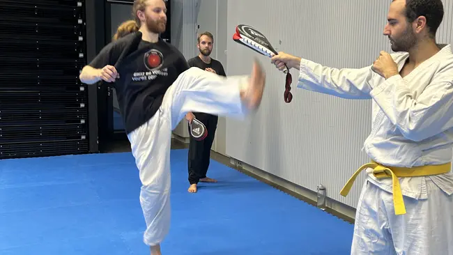 Ein Kampfsportler hebt das Bein zu einem Kick an, während ein Trainer ein Ziel hält. Im Hintergrund sind weitere Personen in der Trainingsumgebung zu sehen. Beide Sportler tragen Trainingsausrüstung, und der Boden ist mit einer blauen Matte ausgelegt.