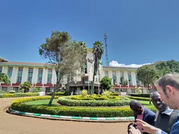 Ein Gruppenfoto vor einem imposanten Regierungsgebäude mit grünen Gärten und Pflanzen. Die Personen im Vordergrund diskutieren, während im Hintergrund die Architektur des Gebäudes und ein klarer blauer Himmel sichtbar sind.