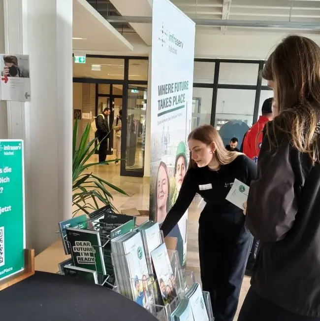 Eine junge Frau steht an einem Stehtisch und schaut sich Flyer und Broschüren an. Im Hintergrund sieht man ein Banner mit dem Text „Unsere Zukunft findet hier statt“. Die Umgebung wirkt modern und einladend.