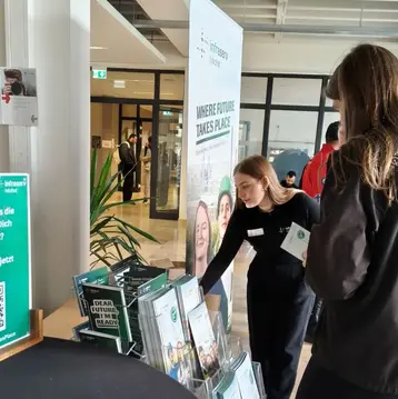 Eine junge Frau steht an einem Stehtisch und schaut sich Flyer und Broschüren an. Im Hintergrund sieht man ein Banner mit dem Text „Unsere Zukunft findet hier statt“. Die Umgebung wirkt modern und einladend.