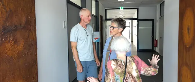 Drei Personen stehen in einem modernen Flur. Eine ältere Frau sitzt in einem Rollator und schaut zu zwei Personen, die miteinander sprechen. Der Mann trägt ein helles T-Shirt, die Frau hat Brille und angelt mit der Hand. Der Flur ist hell erleuchtet und hat graue Fliesen.