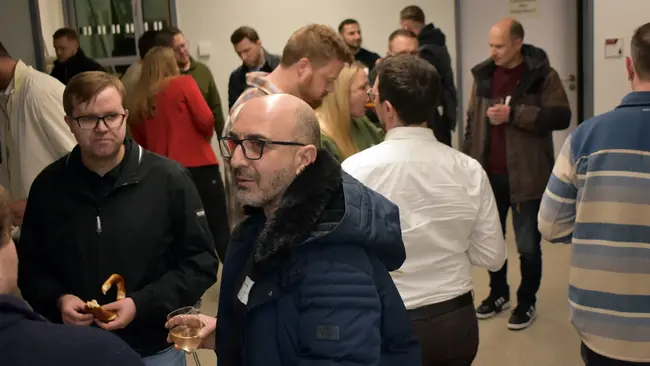 Eine Gruppe von Menschen steht in einem Raum zusammen, spricht und hält Getränke in der Hand. Einige haben Snacks und tragen Alltagskleidung. Die Atmosphäre wirkt gesellig und ungezwungen. Es sind sowohl Männer als auch Frauen anwesend.
