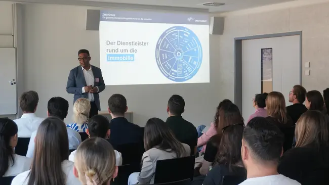 Ein Referent steht in einem Konferenzraum vor einer Gruppe von Zuhörern. Auf einer Leinwand wird eine Grafik mit dem Titel „Der Dienstleister rund um die Immobilie“ gezeigt. Die Zuhörer sind aufmerksam und in verschiedenen Sitzpositionen angeordnet.