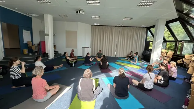 In einem hellen Raum sitzen etwa 15 Personen auf Yogamatten. Sie arbeiten gemeinsam in einer Gruppenübung. Einige blicken zum Lehrer in der Mitte, während andere entspannt auf den Matten sitzen. Durch große Fenster strömt Tageslicht in den Raum.