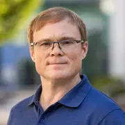 Ein Mann mit Brille und kurzem, blondem Haar steht in einem urbanen Umfeld. Er trägt ein dunkelblaues Polohemd und schaut direkt in die Kamera. Im Hintergrund sind unscharfe Elemente einer modernen Architektur zu sehen.