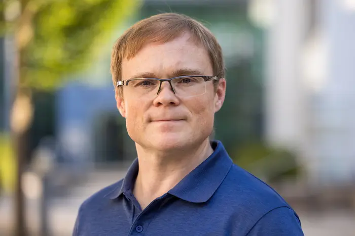 Ein Mann mit Brille und kurzem, blondem Haar steht in einem urbanen Umfeld. Er trägt ein dunkelblaues Polohemd und schaut direkt in die Kamera. Im Hintergrund sind unscharfe Elemente einer modernen Architektur zu sehen.