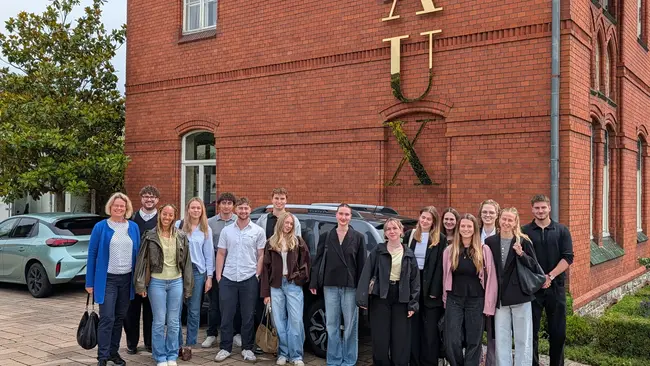 Eine Gruppe von etwa 20 Personen steht vor einem roten Backsteingebäude mit dem großen Schriftzug „VAUX“. Die Personen sind unterschiedlich gekleidet und lächeln in die Kamera. Es ist ein bewölkter Tag. Im Hintergrund sind einige Bäume und ein Auto sichtbar.