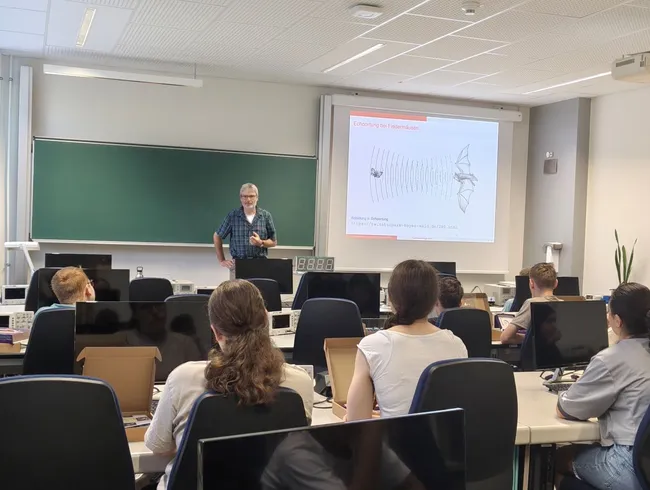 Ein Dozent steht vor einer Gruppe von Studierenden in einem Klassenraum. Eine Folie zeigt ein Diagramm von Fledermäusen und Schallwellen. Die Studierenden sitzen an Computern und hören aufmerksam zu.