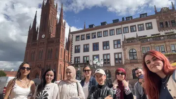 Eine Gruppe von acht Personen steht lächelnd vor einem großen, roten historischen Gebäude mit einer Turmuhr. Im Hintergrund sind moderne Gebäude und ein bewölkter Himmel zu sehen. Die Menschen zeigen eine freundliche, einladende Haltung.