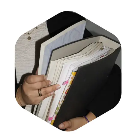 Eine Person hält mehrere Ordner und Dokumente in der Hand. Die Ordner sind teilweise geöffnet und enthalten verschiedene Papiere. Die Person trägt Schmuck und hat eine graue Bluse an.
