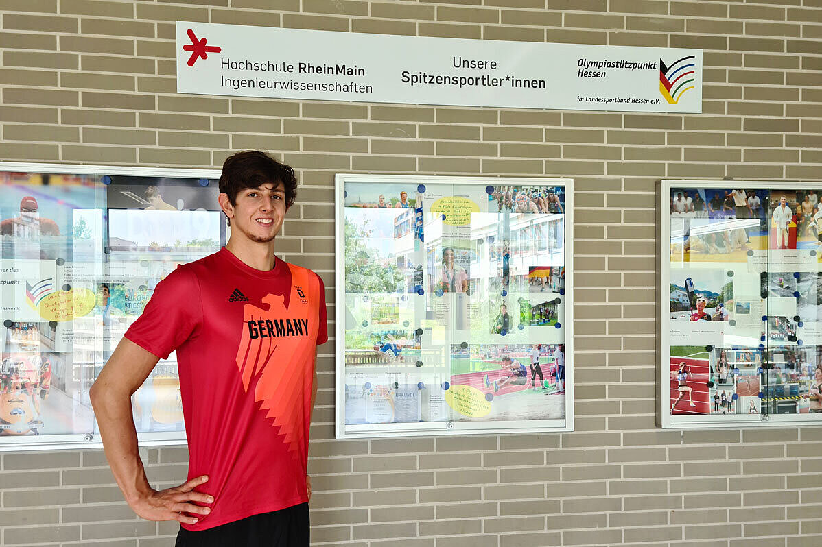 Porträt von Lucas Matzerath, Schwimmen Lucas Matzerath steht auf dem Campus Rüsselsheim vor einem Glaskasten mit Fotos von Spitzensportler:innen der Hochschule RheinMain.