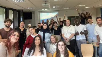 Eine Gruppe von etwa 30 Menschen steht in einem Raum und lächelt. Einige Personen heben die Hände. Sie sind vielfältig gekleidet und scheinen Freude zu haben. Im Hintergrund ist ein Projektor mit einer Präsentation zu sehen.