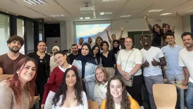 Eine Gruppe von etwa 30 Menschen steht in einem Raum und lächelt. Einige Personen heben die Hände. Sie sind vielfältig gekleidet und scheinen Freude zu haben. Im Hintergrund ist ein Projektor mit einer Präsentation zu sehen.