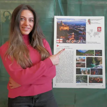 Eine Frau mit langen Haaren trägt einen rosa Pullover und zeigt auf ein Plakat, das Informationen über Georgien enthält. Das Plakat zeigt Fotos von Landschaften und Sehenswürdigkeiten sowie einen Text über das Land.