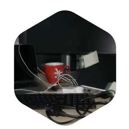 Ein Arbeitsplatz mit einem Laptop und einer Tastatur. In der Mitte steht eine rote Tasse, umgeben von mehreren Kabeln. Im Hintergrund ist ein Notizzettel zu sehen. Der Fokus liegt auf dem chaotischen Kabelsalat und dem Getränk.