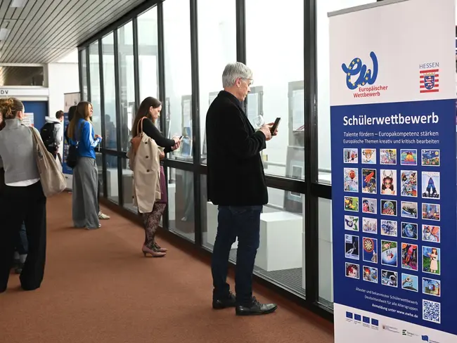 Eine Gruppe von Menschen steht in einem Raum und schaut siche ein Ausstellung an, einige betrachten  die Wand mit einer Infotafel zum Schülerwettbewerb beobachten. Die Tafel zeigt verschiedene Bilder und Informationen zu den Themen des Wettbewerbs.