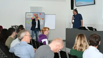 Ein Publikum sitzt in einem Seminarraum, während zwei Männer auf einer Bühne sprechen. Der Mann links hält ein Mikrofon und spricht, der andere steht neben ihm mit einem Mikrofon in der Hand. Im Hintergrund sind Flipcharts und ein Präsentationsbildschirm sichtbar.