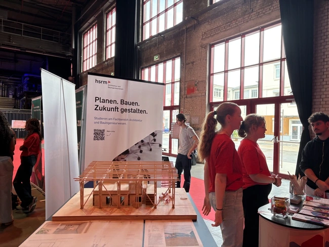 Ein Messestand mit zwei Personen in roten T-Shirts, die an einem Modell eines Gebäudes arbeiten. Im Hintergrund steht ein Banner mit der Aufschrift „Planen. Bauen. Zukunft gestalten.“ Der Stand präsentiert Informationen über Studienangebote im Bereich Architektur und Bauingenieurwesen.