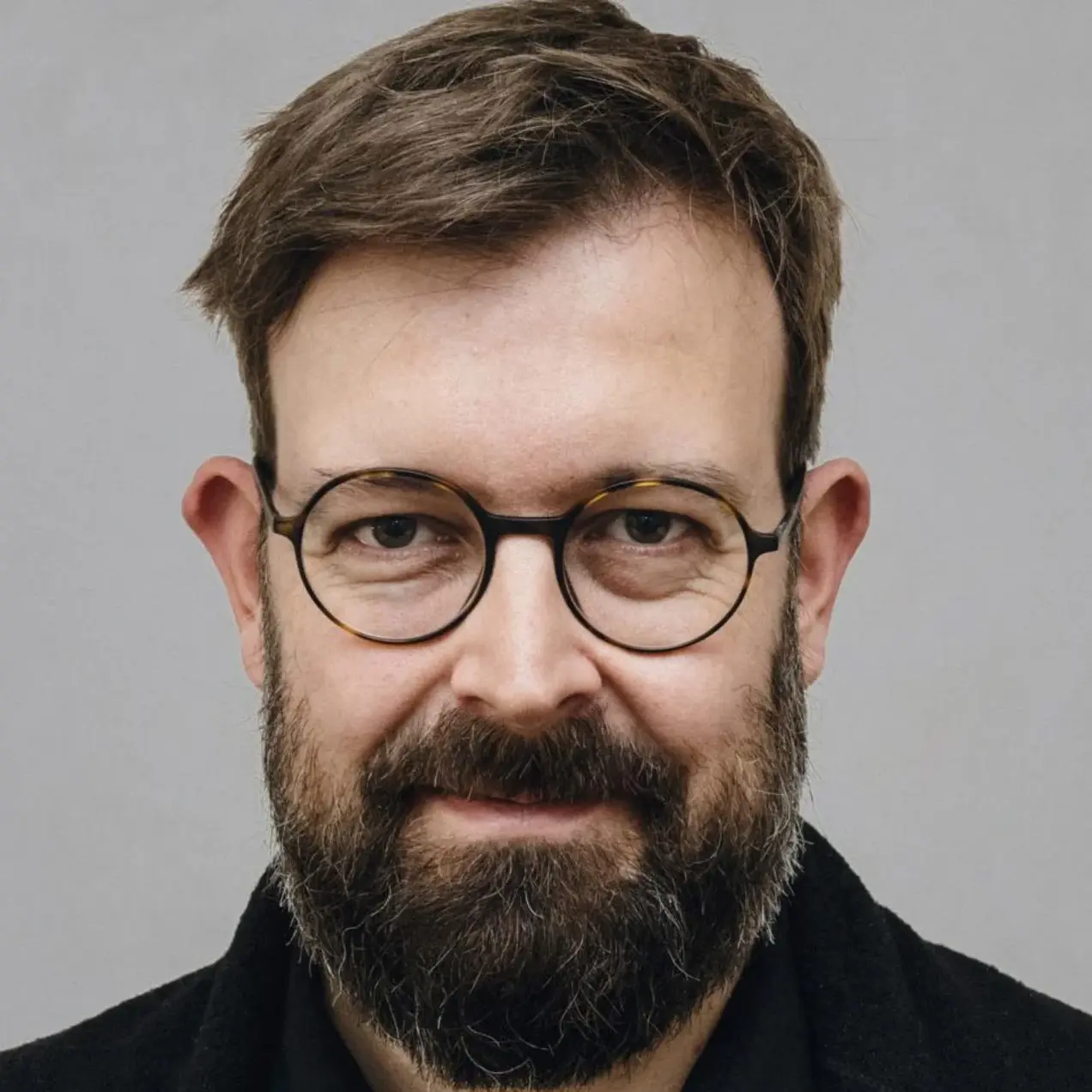 Porträt eines Mannes mit Brille und einem dichten, gepflegten Bart. Er trägt ein schwarzes Oberteil und hat eine freundliche Ausstrahlung. Der Hintergrund ist neutral und hell.