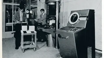 Ein Baustofflabor mit zwei Männern. Einer steht an einem Tisch mit Proben, während der andere an einer Prüfstation arbeitet. Im Hintergrund ist ein Gerät zur Prüfung von Beton zu sehen, das mit einer großen Uhr ausgestattet ist.