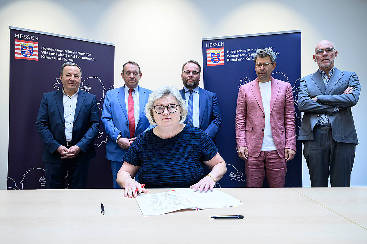 Prof Dr. Eva Waller sitzt an einem Tisch, auf dem der Hochschulpakt liegt. Hinter ihr stehen Prof. Dr. Karim Khakzar, Prof. Dr. R. Alexander Lorz, Timon Gremmels, Prof. Dr. Thomas Nauss, Prof. Elmar Fulda 