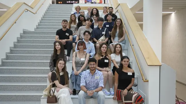 Eine Gruppe von etwa 20 Personen sitzt und steht auf einer Treppe in einem modernen Gebäude. Die Menschen sind vielfältig gekleidet und lächeln zur Kamera. Auf einigen von ihnen sind Namensschilder zu sehen. Die Atmosphäre ist freundlich und einladend.