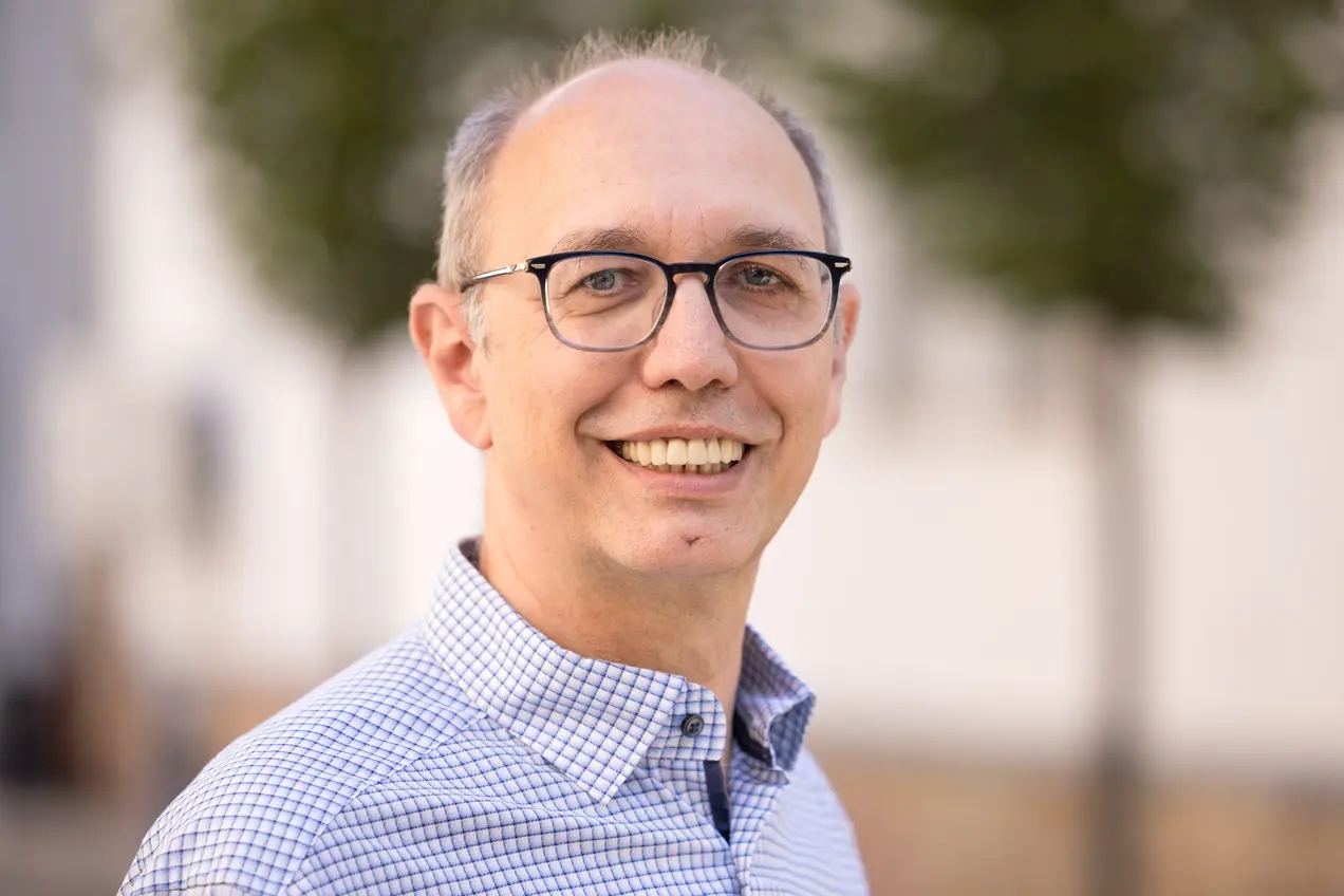 Ein lächelnder Mann mit Brille steht vor einem unscharfen Hintergrund. Er trägt ein kariertes Hemd und hat graues, kurzes Haar. Die Stimmung wirkt freundlich und einladend.